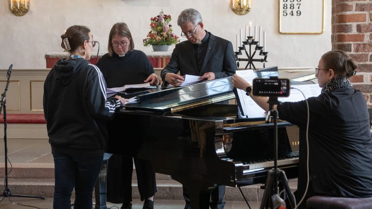 En grupp personer står vid ett flygel i en kyrksal och ser på papper.