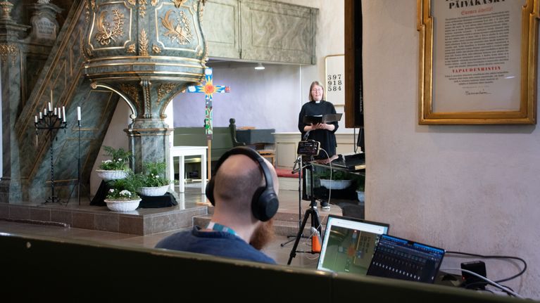 En person arbetar vid en dator i en kyrka medan en präst står framför en predikstol och sjunger.