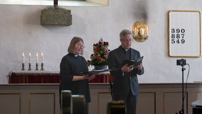 Två präster står framför ett altare i en kyrka och håller böcker i handen.