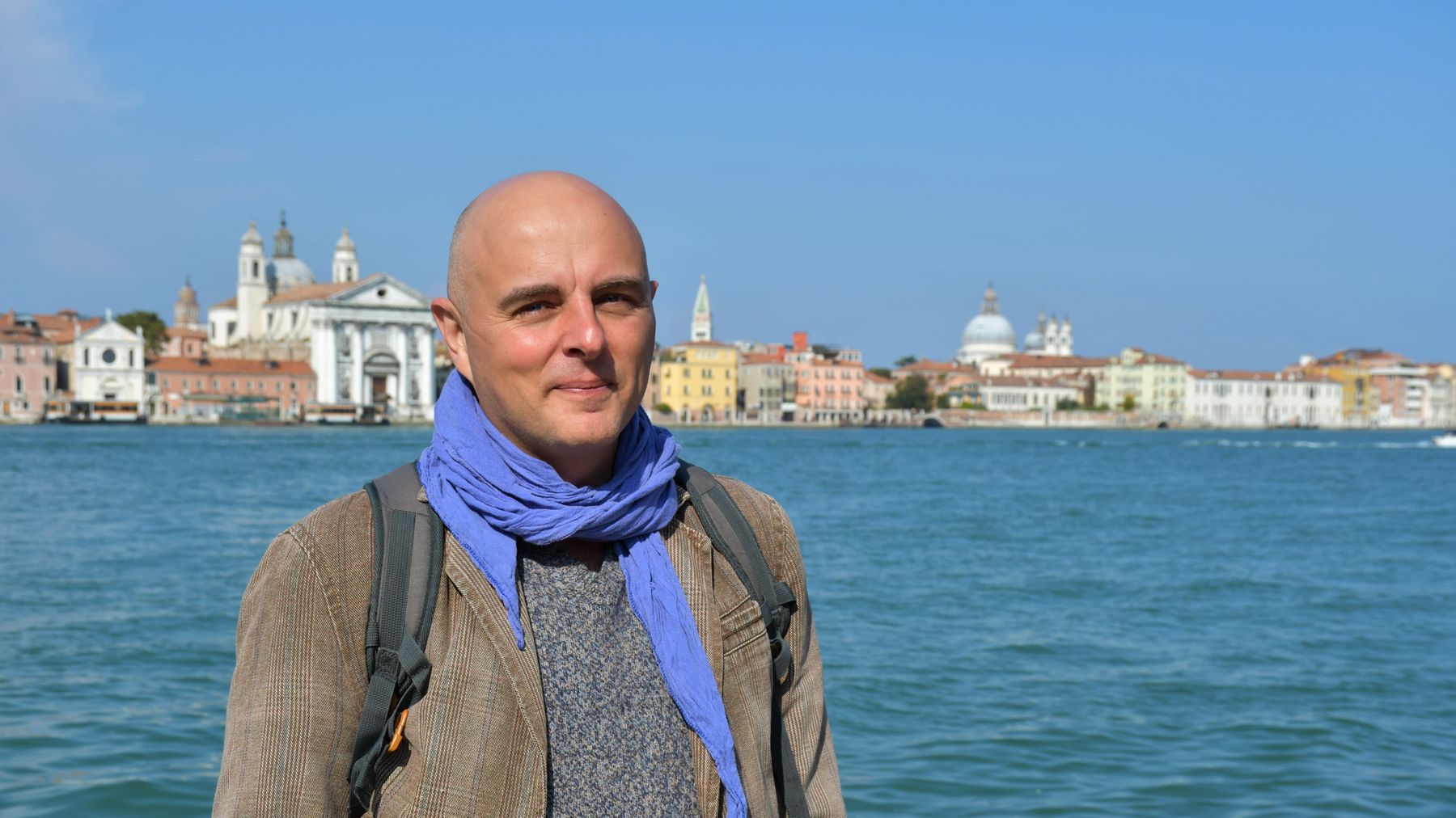 En man utan hår i brun jakca med blå scarf tittar in kameran och ler. I bakgrunden azurblått hav med Venedig i silhuett i bakgrunden.