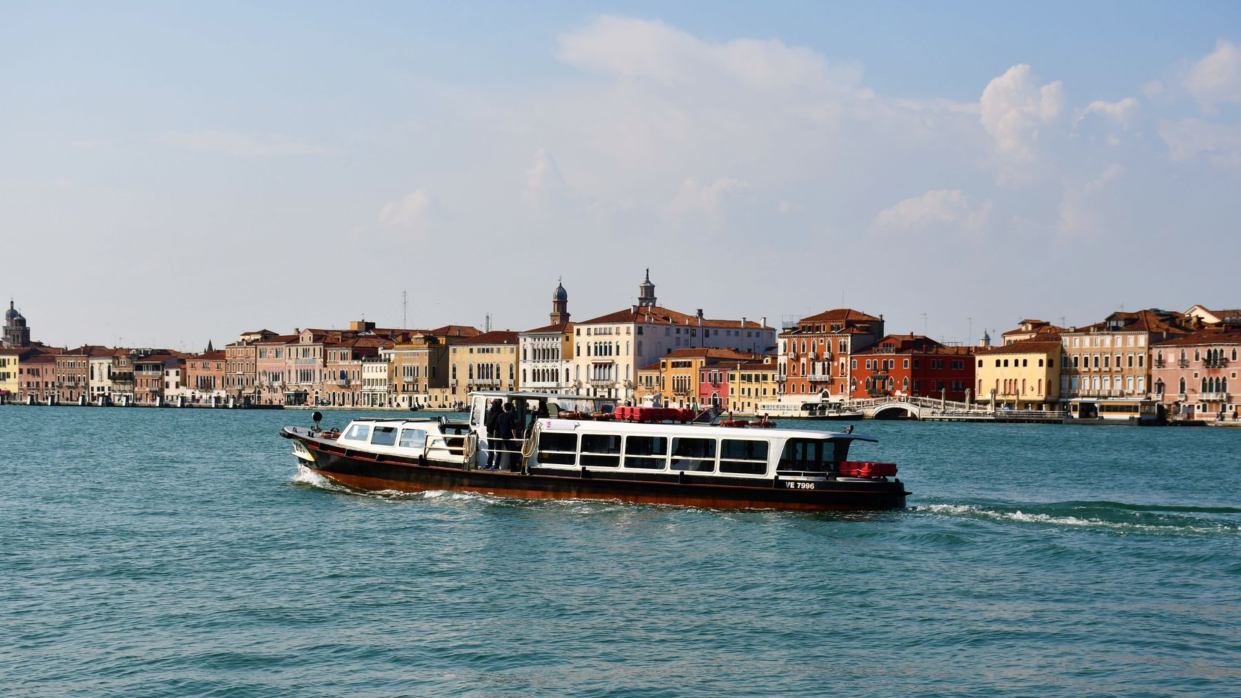 En vattenbuss åker på ett azurblått hav med Venedig i silhuett i bakgrunden.