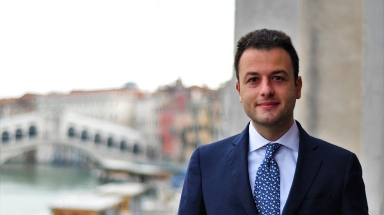 Man med mörkt hår i mör kostym står framför en stadsvy i Venedig.