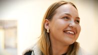 En person med blont hår, klädd i en jacka med ullfodrad krage, har guldfärgade örhängen och ett halsband. Bakgrunden visar en ljus vägg med en tavla.