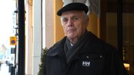 En äldre man med hatt och jacka står vid en husvägg.