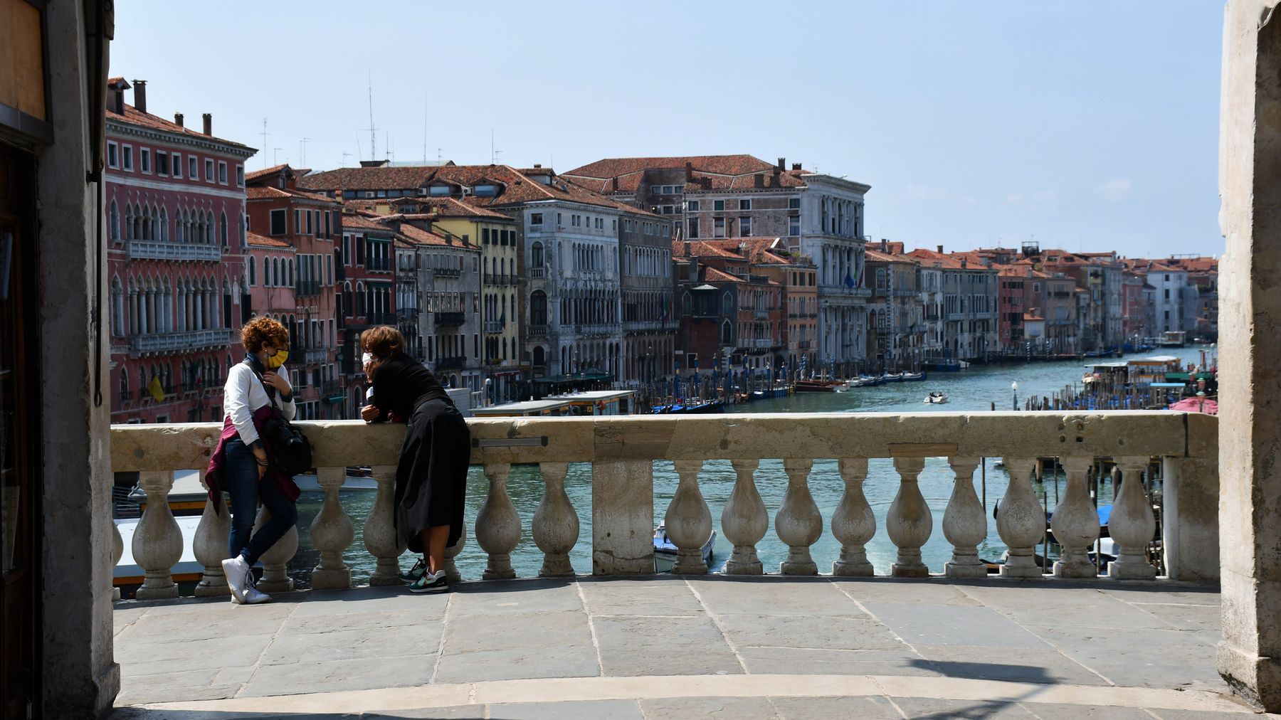 Två kvinnor med munskydd står lutade mot varandra och mot ett broräcke i vit sten. Bakom räcket syns en kanal i Venedig och husen på bägge sidor.