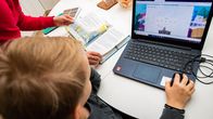 A boy seen from above working on a laptop while an adult stands off to the left holding an open textbook, with neither face visible.