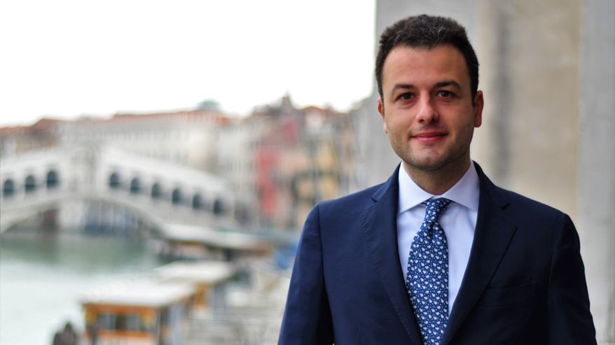 En man med kort mörkt hår, mörk kostym, ljusblå skjorta och vitprickig blå kravatt ler mot kameran. I bakgrunden suddigt en bro i Venedig.