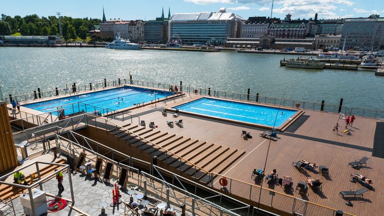 Two swimming pools on a pier by a harbour, with boats and buildings in the background.