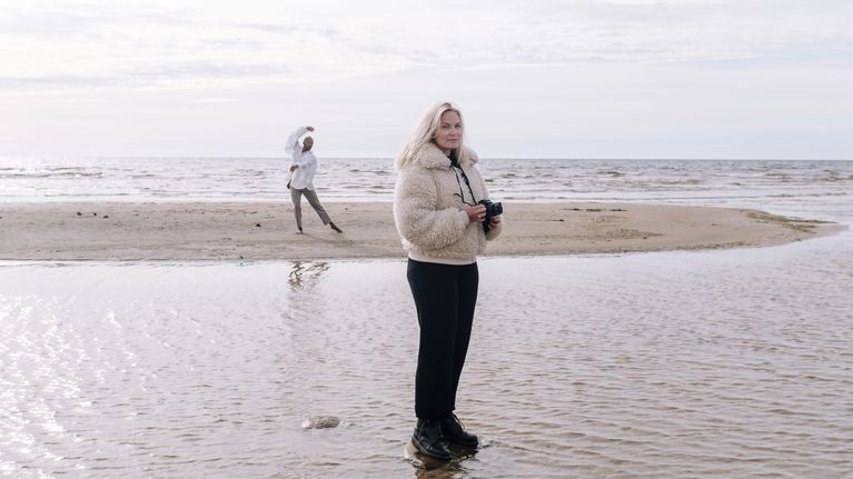 Emilia Hernesniemi ja Jani Toivola Hailuodossa meren rannalla. Toivola tanssii taustalla valkoisessa paidassa ja Hernesniemi seisoo etualalla kamera käsissään.