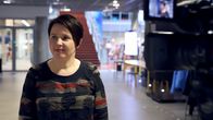 Tummahiuksinen nainen rakennuksen aulassa, taustalla portaikko ja etualalla televisiokamera.