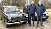 Tre personer står utomhus bredvid en svart bil och en grå bil på en grusväg med träd i bakgrunden.