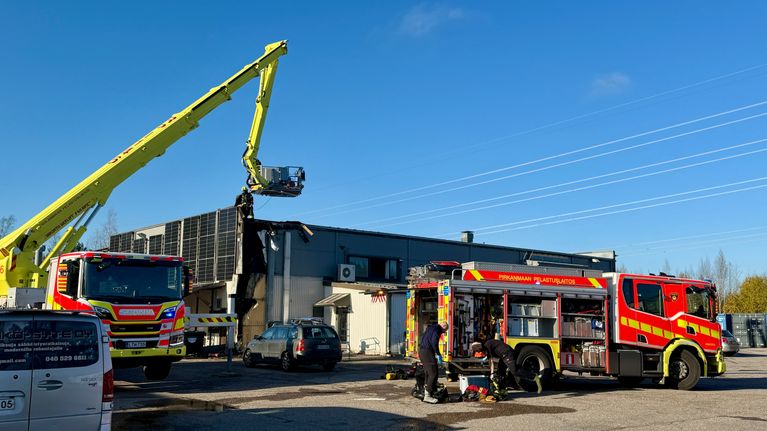 Kaksi paloautoa ja useita palomiehiä työskentelee teollisuushallin pihalla, toinen paloautoista varustettu nostokorilla.