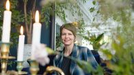 Elisa istuu sohvalla, katsoo kameraan ja hymyilee. Kuvattu kukka-asetelman takaa.