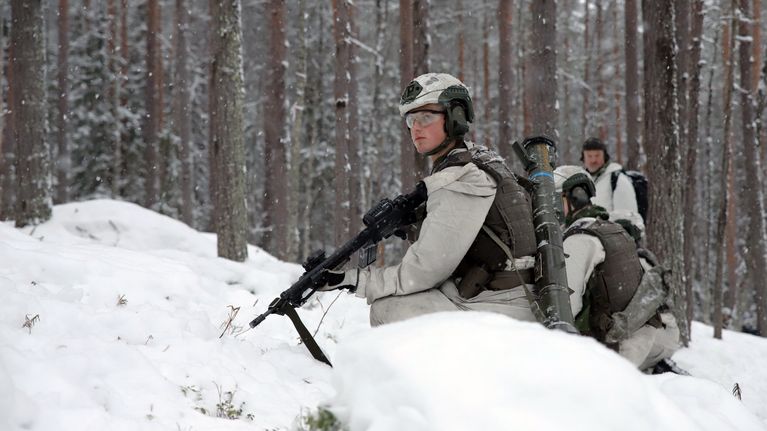 Soldater tränar i skogen under vinterförhållanden, klädda i kamouflage för snömiljöer och utrustade med vapen och kommunikationsutrustning.