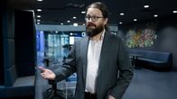 Otso Kivekäs, klädd i en grå kavaj och vit skjorta, står i en halvfigursbild och gestikulerar med händerna medan han talar till journalisten. I bakgrunden syns ett foajéliknande utrymme.