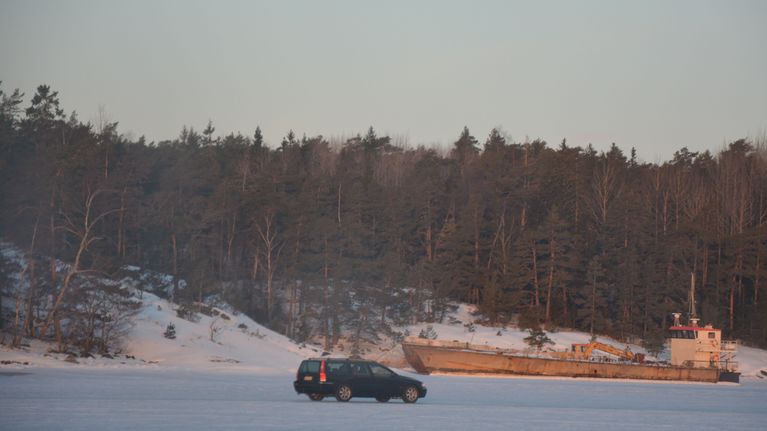 Auto ajaa jäälllä, taustalla näkyy rannan tuntumassa oleva kuljetusalus.