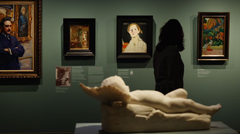 En person står framför flera tavlor på en grön vägg i ett konstmuseum; en liggande staty i förgrunden.