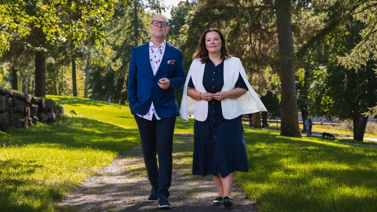 Två personer går sida vid sida på en grusväg i en park med gröna träd och gräsmatta.