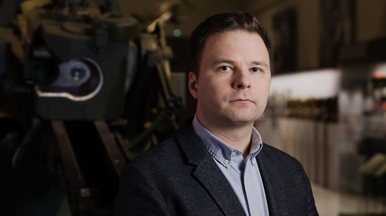 Kustannustoimittaja Aleski Pöyry seisoo ilmatorjuntatykin edessä sotamuseossa, puolilähikuva.