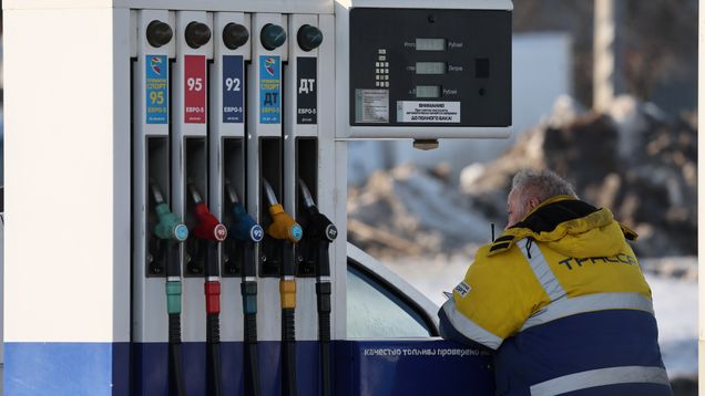 Ihminen kelta sinisessä työtakissa nojaa polttoaine pumppuun huoltoasemalla.