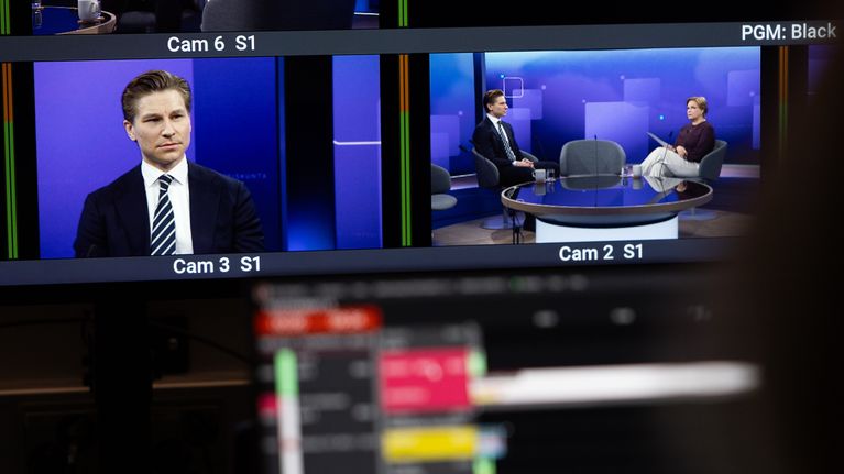 Video screens in a TV studio, one screen showing a man on camera in a dark suit, and the other screen shows a journalist sitting beside him.