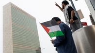 Kaksi miestä seisoo YK:n päämajan edessä, toisella Palestiinan lippua ja toisella palestiinalaishuivi.