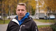 Mies sinisessä hupparissa ja mustassa ulkoilutakissa ulkona, taustalla syksyisiä puita ja pysäköityjä autoja.