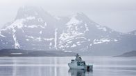 Ett grått militärfartyg glider över stilla vatten med ett storslaget och delvis snötäckt berglandskap i bakgrunden.