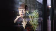 Riikka Leinonen katsoo vakavailmeisenä kameraan ikkunan takaa, etualalla on kukkakimppu.