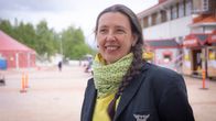 Kuvassa henkilö seisoo ulkoilmassa, pukeutuneena mustaan takkiin, jossa on siivet korostava merkki, sekä keltaiseen huiviin ja huppariin. Taustalla näkyy rakennuksia ja teltta festivaaleilla.