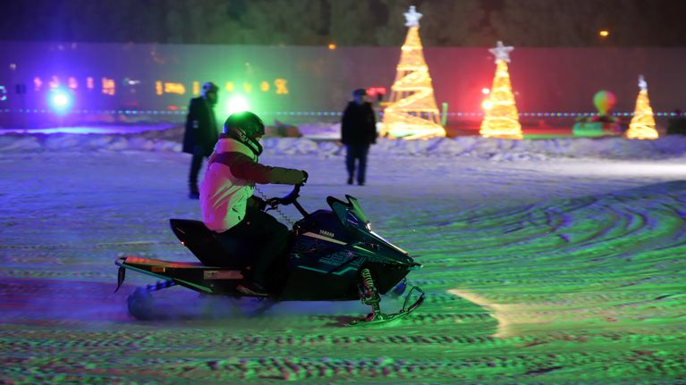 Talvisessa valopuistossa nuori ajaa pientä moottorikelkkaa kahden henkilön valvoessa tilannetta.