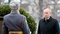 Putin seisoo mustassa päällystakissa, kravatti kaulassaan seppeleen edessä. Etualalla seisoo sotilas harmaassa manttelissa ja karvalakissa ja pitelee seppelettä.