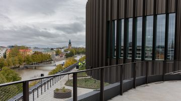 En byggnad med glasfasad bredvid en å, träd och hus syns i bakgrunden under molnig himmel.