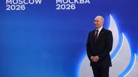 Hymyilevä Vladimir Putin seisoo lavalla mustassa puvussa ja viininpunaisessa kravatissa. Sinisellä seinällä lukee venäjäksi 8. Moskovan huippukokous.
