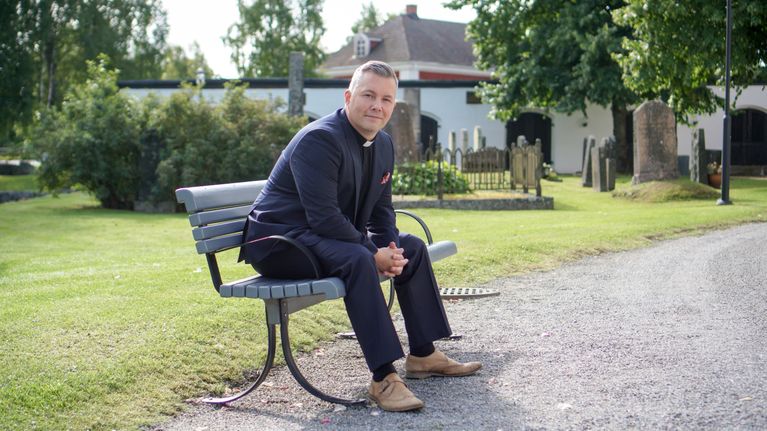 En man i prästskjorta och kostym sitter på en bänk vid en kyrkogård med gravstenar och grönska i bakgrunden.