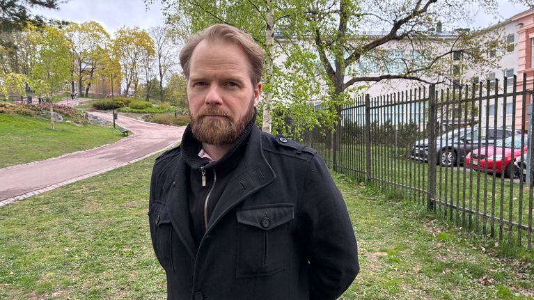 En person med mörk jacka står på en gräsmatta intill ett staket och flera träd.