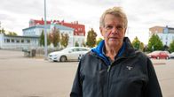 En man står på en parkeringsplats med byggnader i bakgrunden. Han bär en mörk jacka.
