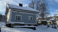Vaaleansininen omakotitalo lumen peittämällä pihalla, taustalla lehdettömiä puita ja sininen taivas.