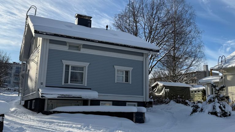 Vaaleansininen omakotitalo lumen peittämällä pihalla, taustalla lehdettömiä puita ja sininen taivas.