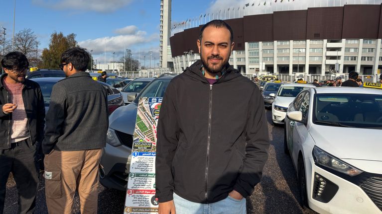 En man står framför parkerade taxibilar och ett högt torn vid en stor arena, med flera människor i bakgrunden.