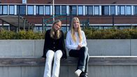 Två unga personer sitter på en träbänk utomhus framför en skolbyggnad med stora fönster och en klocka på fasaden.