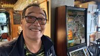 En person står bredvid en spelautomat i en restaurang eller bar. Automaten har olika färgglada symboler och skärmar. I taket hänger glas, och i bakgrunden syns träpaneler och bord.