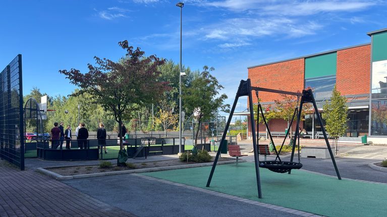 Flera personer står på en inhägnad fotbollsplan intill en lekplats med gunga utanför en tegelbyggnad.