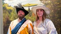 Historiallisiin asuihin pukeutuneet mies ja nainen. Taustalla kuva jokimaisemasta.