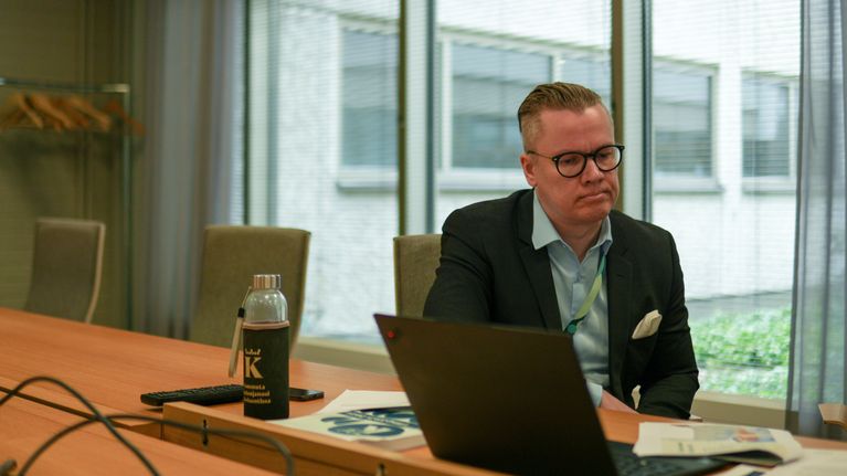 En man sitter vid ett konferensbord med en bärbar dator, papper och en vattenflaska framför sig.
