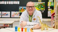 Mies hymyilee pöydän ääressä, jolla seisoo dinosauruslelu ja värikkäitä rakennuspalikoita.
