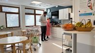 Nainen tekee sairaala-asussa keittiöaskareita, mies normaalissa vaatetuksessa vieressä. Vauva on pyörillä olevassa kaukalossa.