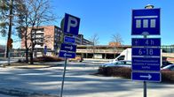 Pysäköintiä ohjaavia liikennemerkkejä pysäköintialueella. Taustalla iso rakennus ja siinä välissä muutamia autoja parkkipaikalla.