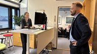 Nainen seisoo tietokoneen äärellä ja keskustelee miehen kanssa toimistohuoneessa, taustalla istuu kaksi henkilöä.