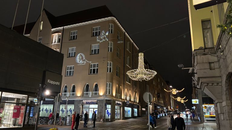 Kadulla kävelee ihmisiä. Kadun yllä on jouluvaloja ja rakennuksen seinään on heijastettu muumihahmoja.